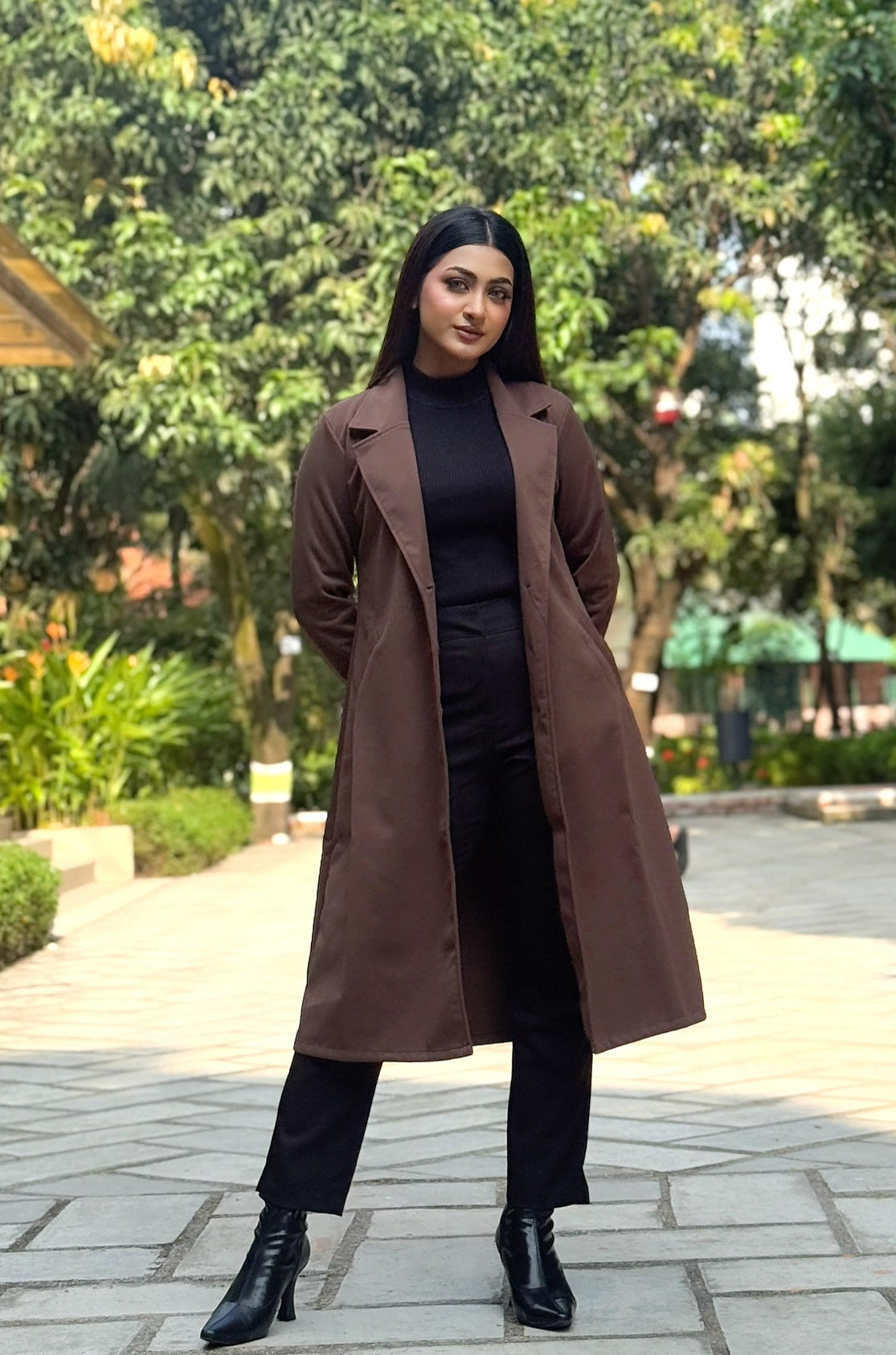 Woman in a brown coat standing in a park with trees and buildings in the background