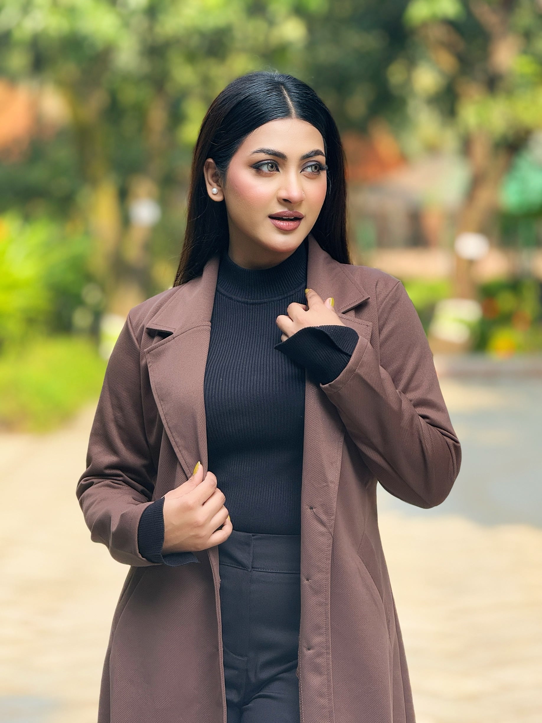 Woman wearing a brown coat and black outfit outdoors with greenery in the background