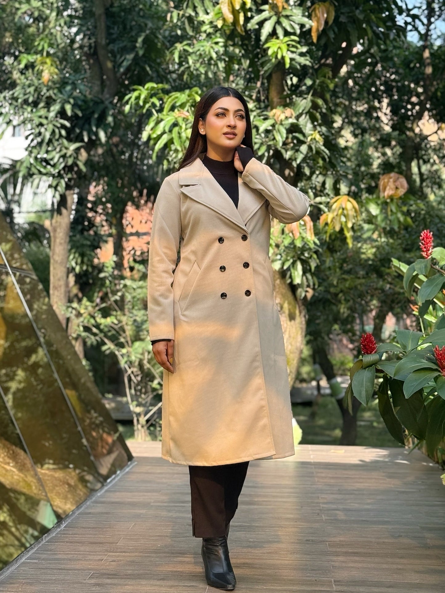 Woman in a beige trench coat walking on a wooden bridge with greenery in the background
