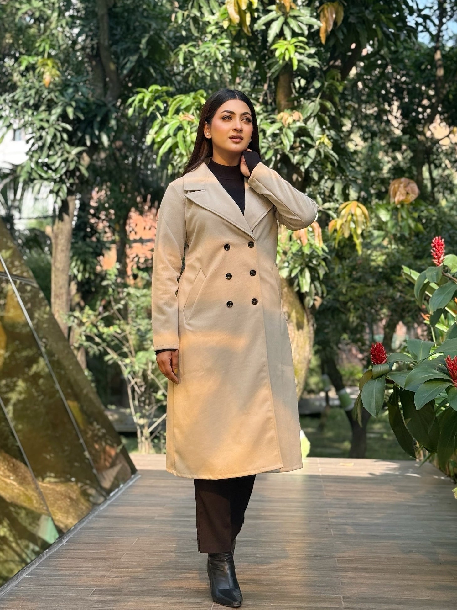 Woman in a beige trench coat walking on a wooden bridge with greenery in the background