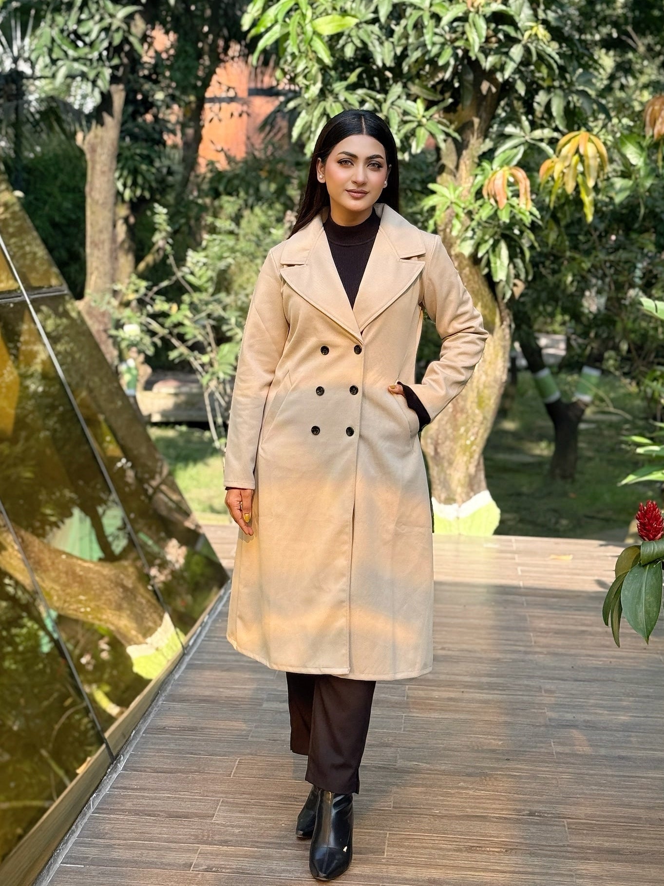 Woman in a beige trench coat standing on a wooden path with greenery around
