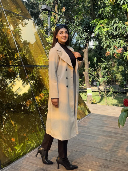 Woman in a long coat standing in front of a reflective surface with greenery in the background