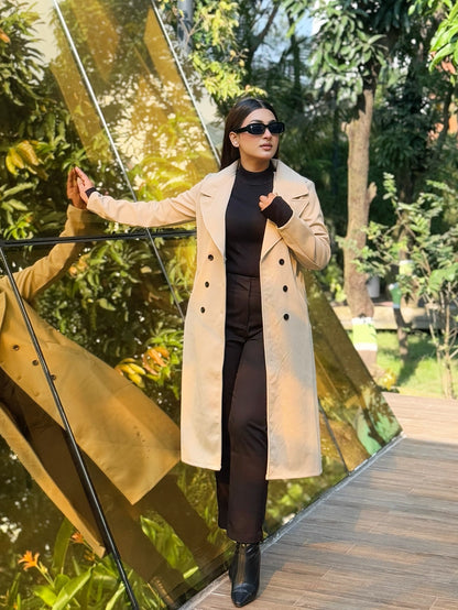 Woman in a beige coat standing in front of a mirrored sculpture with greenery in the background