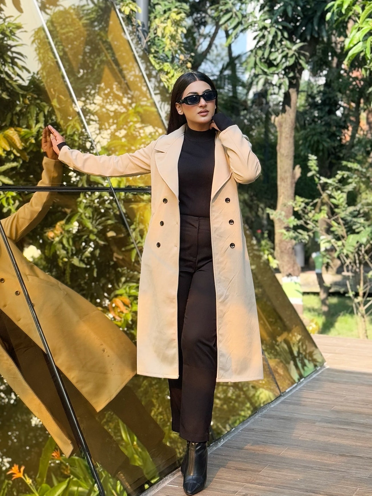 Woman in a beige coat standing in front of a reflective gold surface with trees in the background