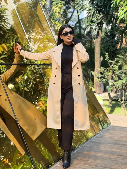 Woman in a beige coat standing in front of a reflective gold surface with trees in the background