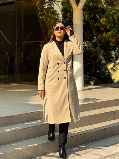 Woman in a beige coat standing on steps outdoors with modern architecture in the background