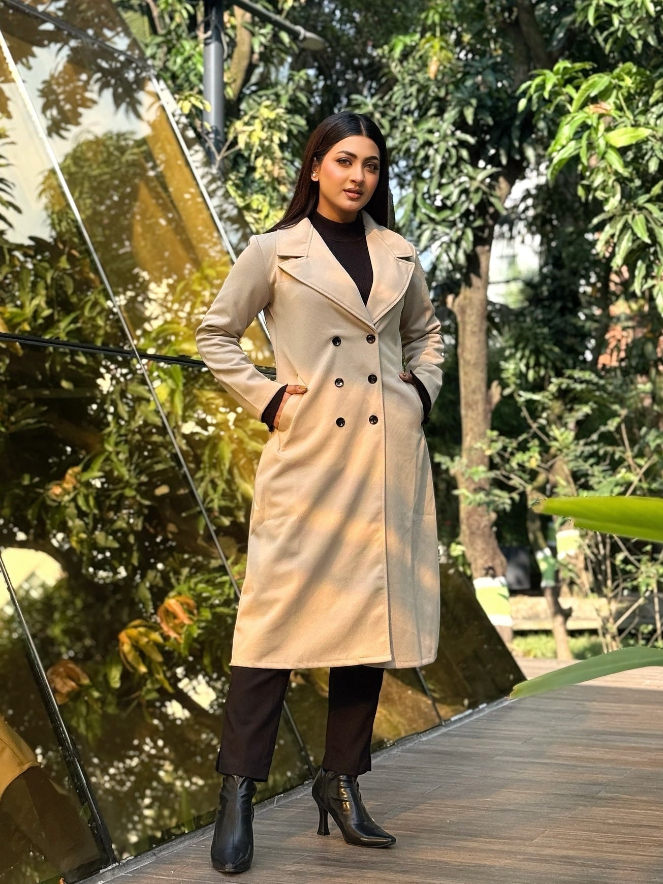 Woman in a beige trench coat standing outdoors with greenery in the background