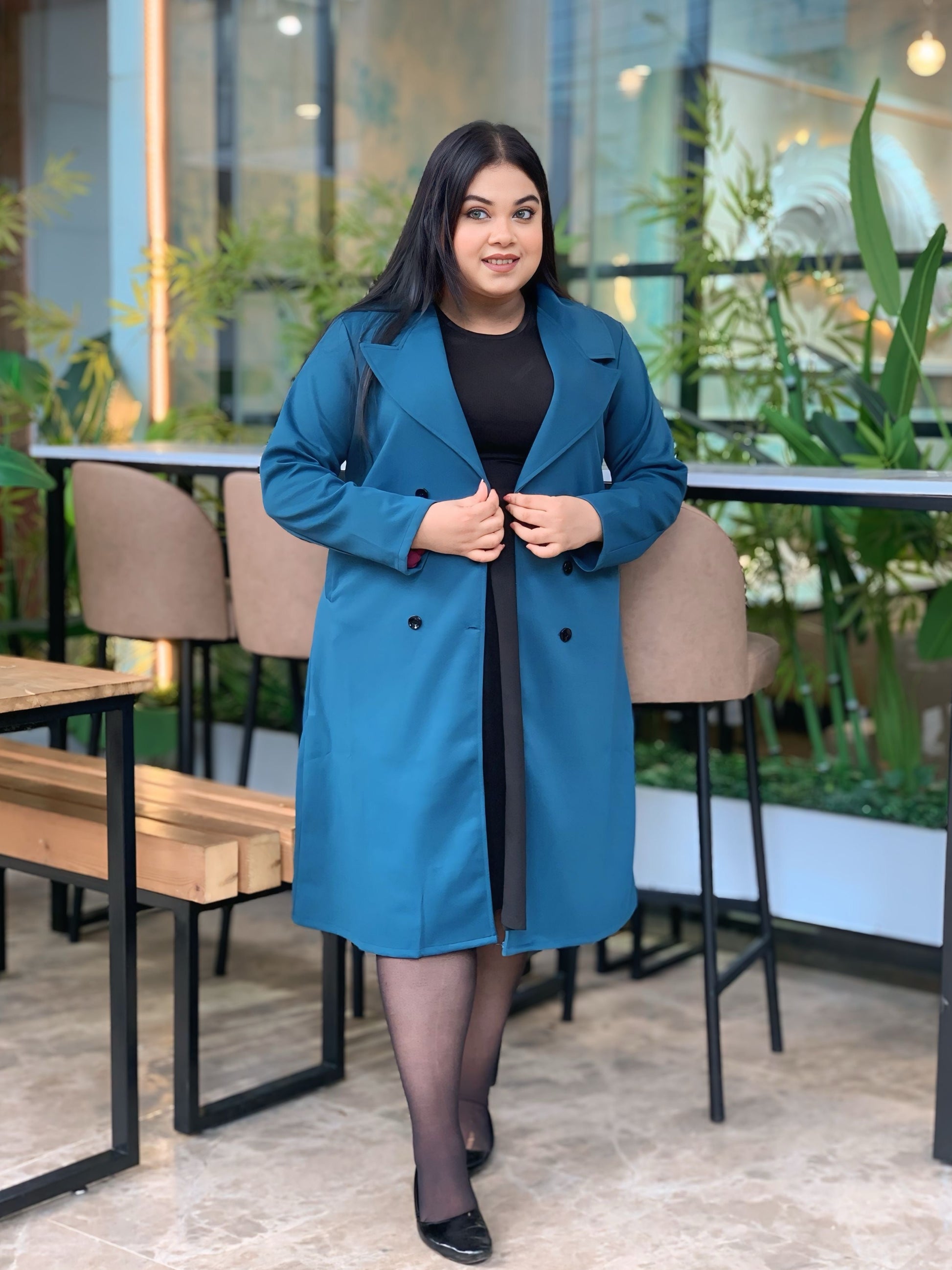 Woman wearing a blue coat in an indoor setting with plants and furniture.