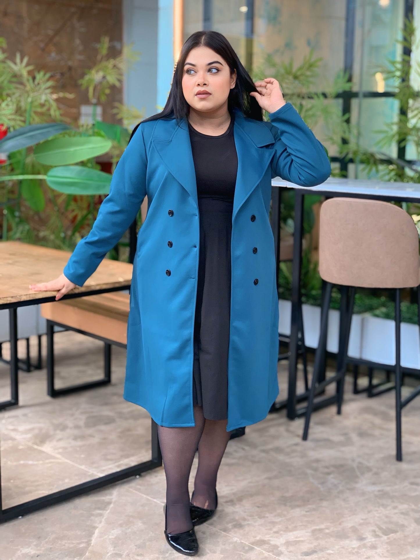 Woman wearing a blue coat in an outdoor setting with plants and tables.