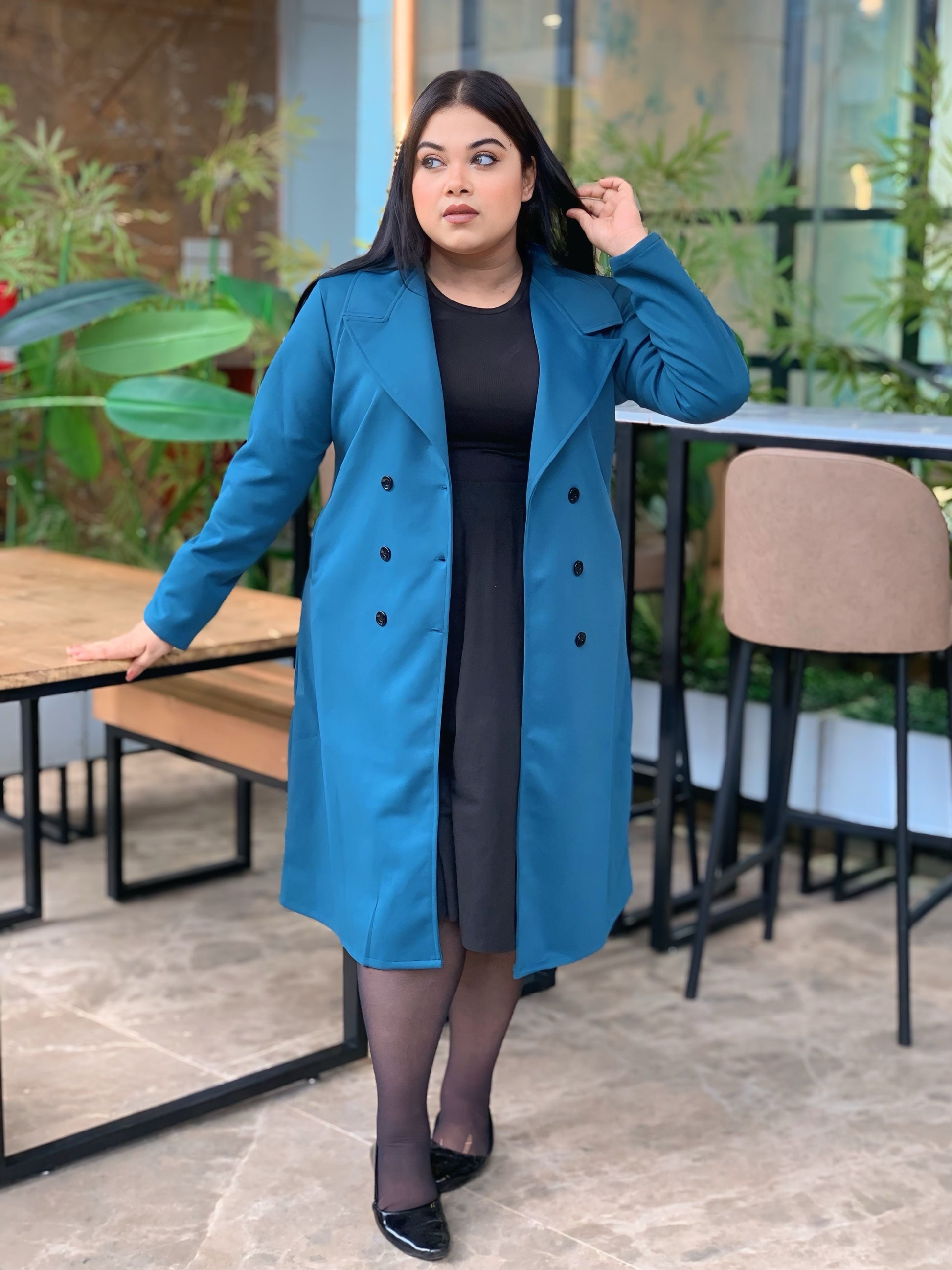 Woman wearing a blue coat in an outdoor setting with plants and tables.