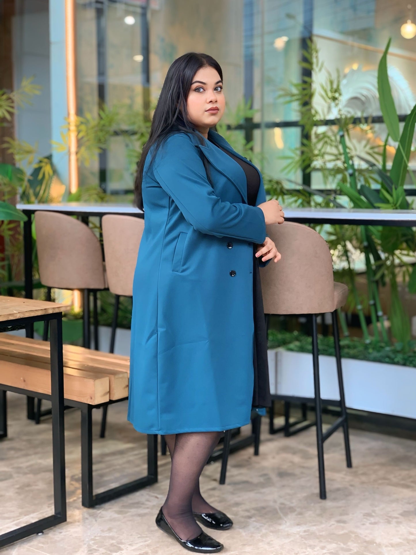 Woman in a blue coat standing in an indoor setting with plants and tables.