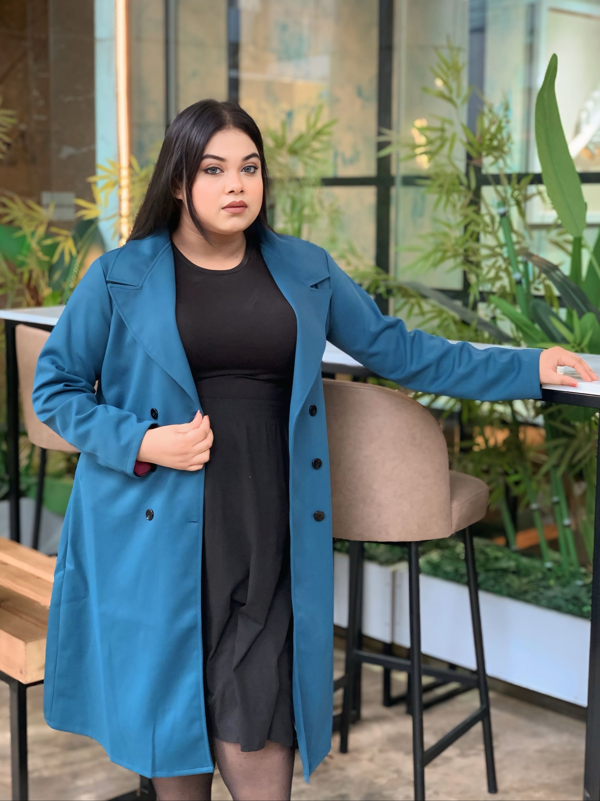 Woman wearing a blue coat over a black dress in an indoor setting with plants.