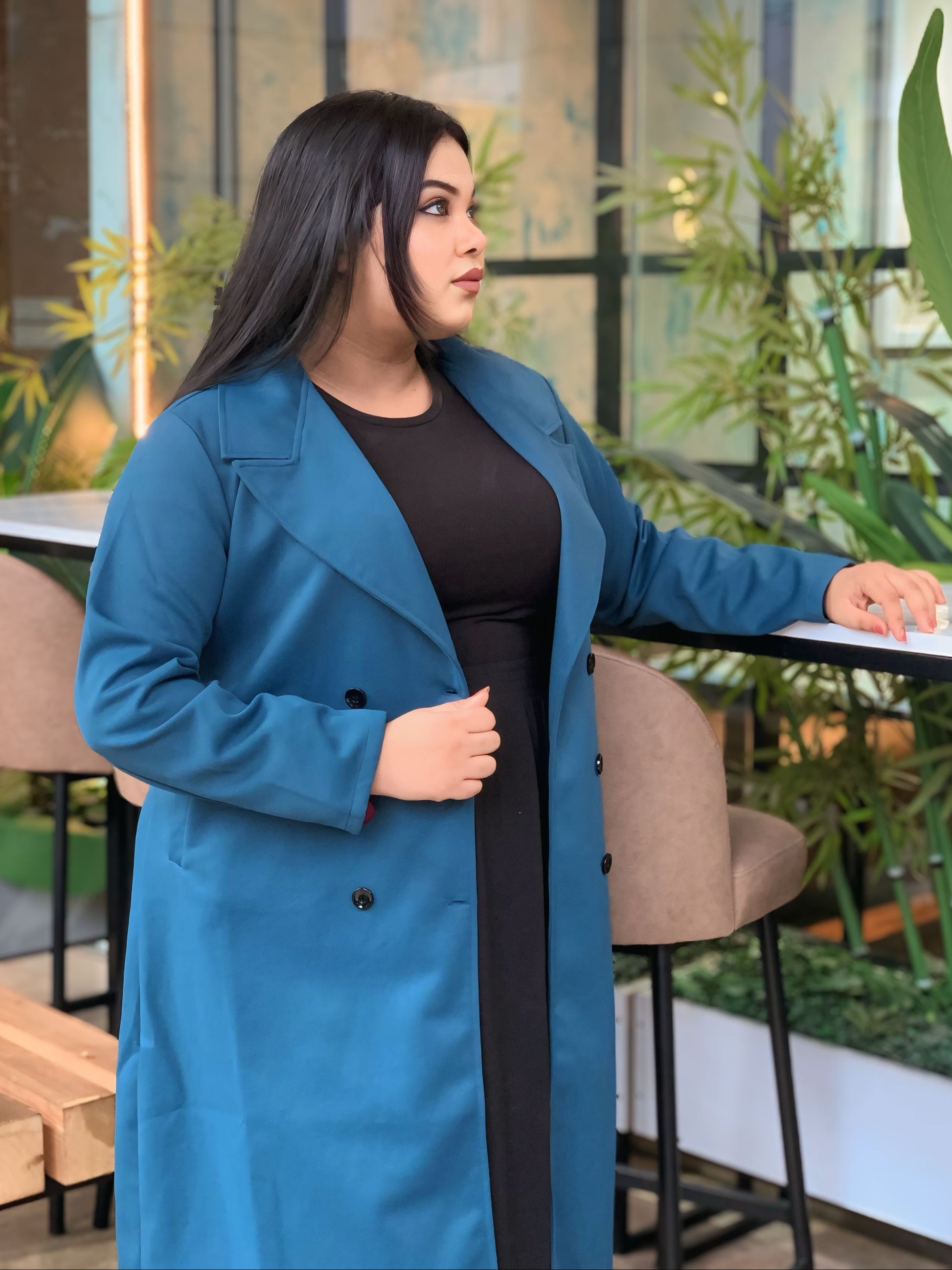 Woman wearing a blue coat in an indoor setting with plants and furniture.