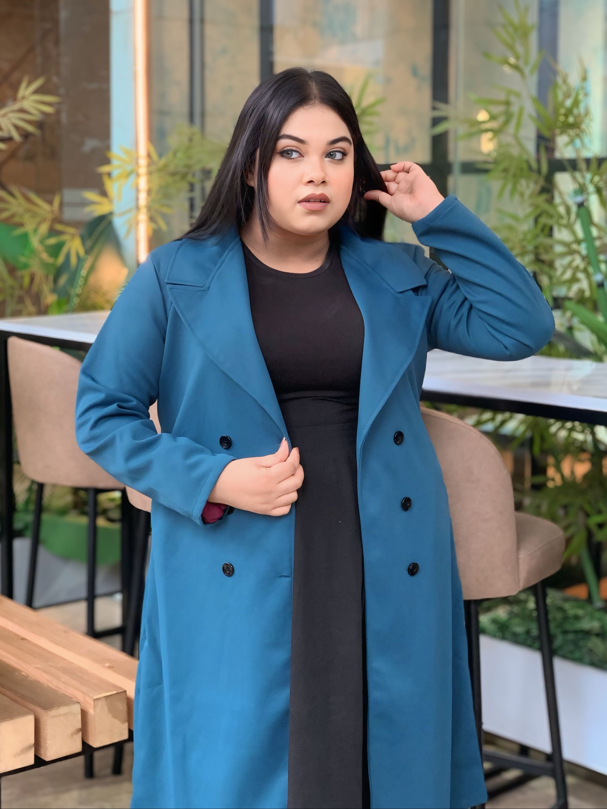 Woman wearing a blue coat in an outdoor setting with plants and tables.
