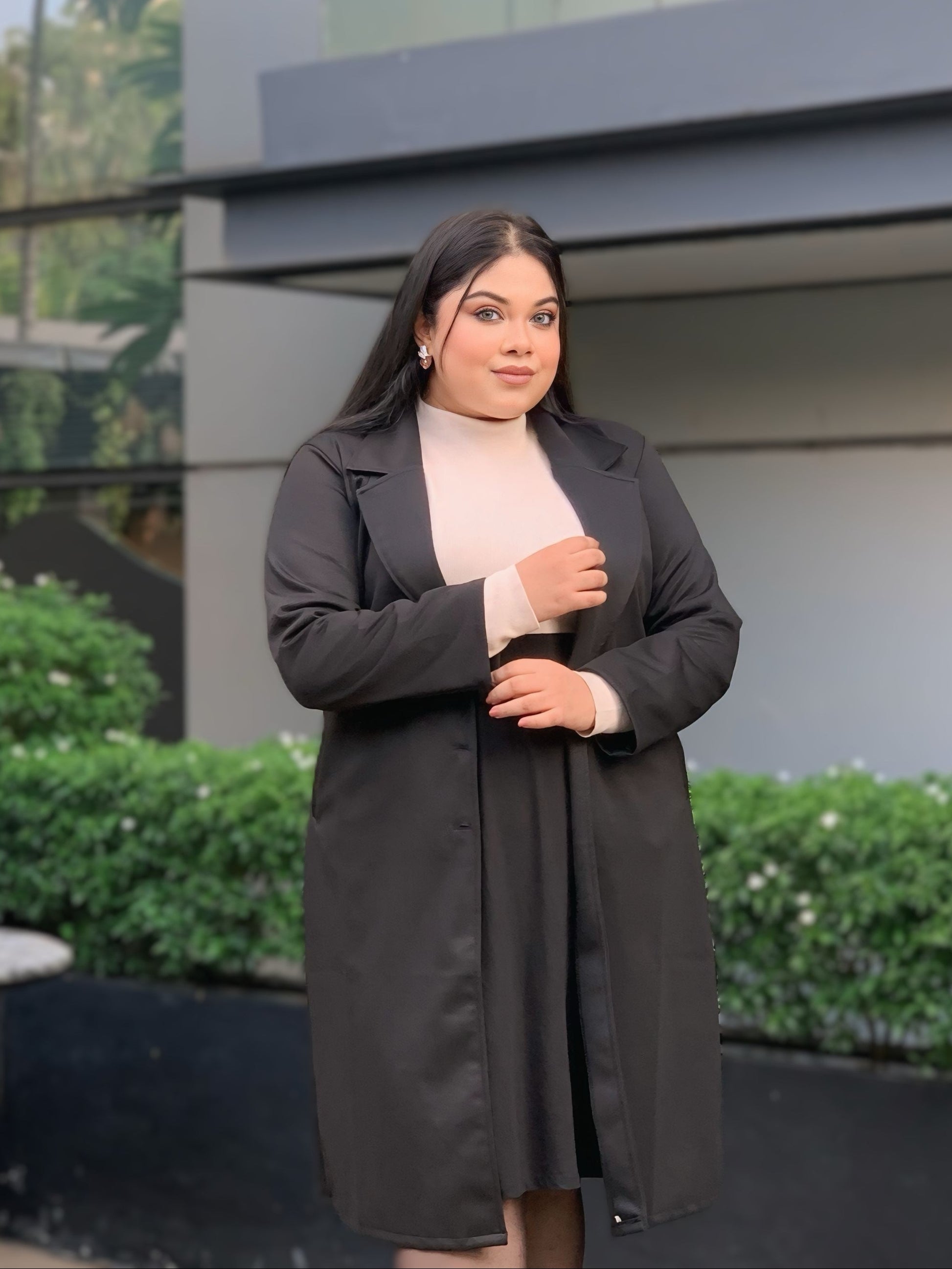 Woman in a black coat standing outdoors with a building and greenery in the background