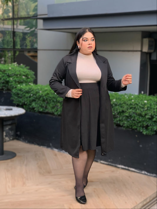 Woman in a black coat and skirt standing outdoors with greenery and a building in the background