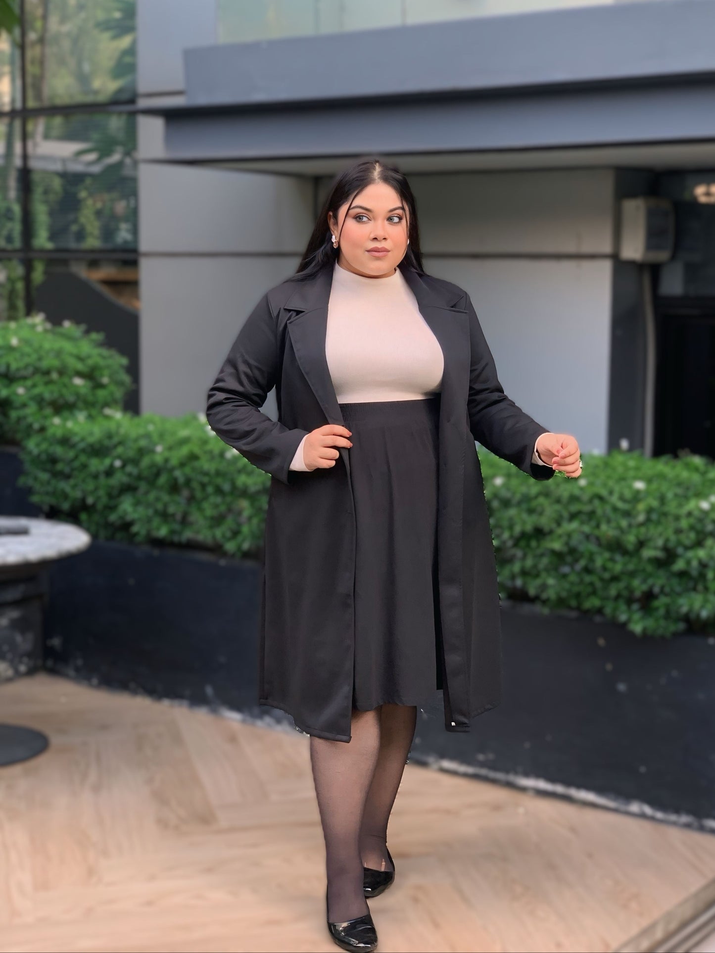 Woman in a black coat and skirt standing outdoors with greenery and a building in the background