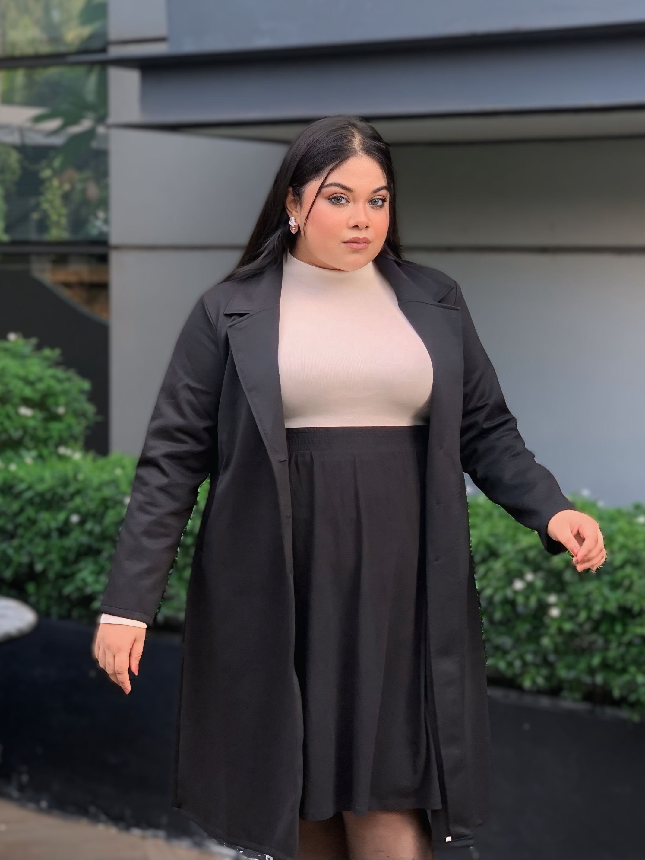 Woman wearing a black coat and skirt standing outdoors with greenery in the background