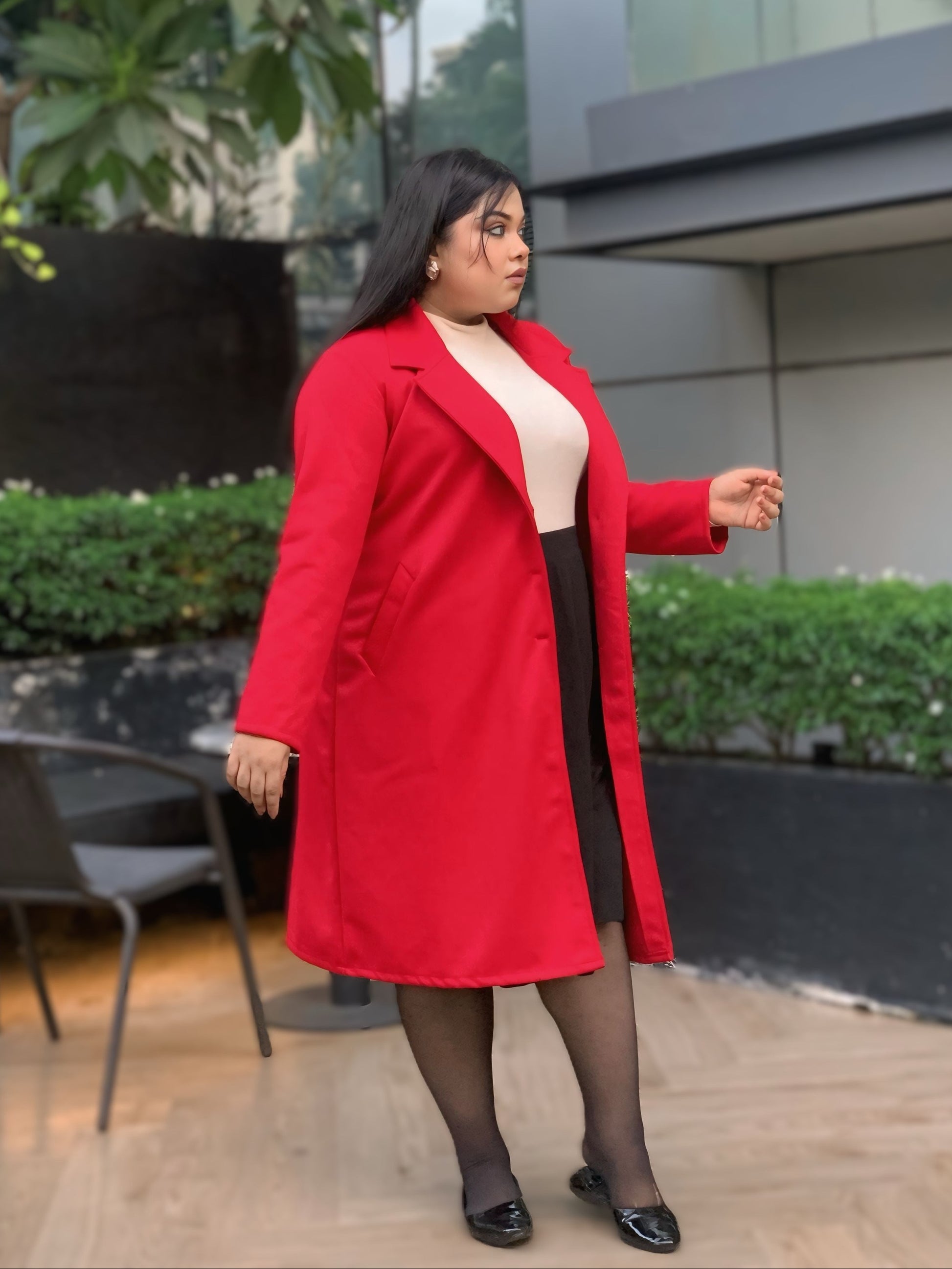 Woman wearing a red coat standing outdoors with greenery and a building in the background