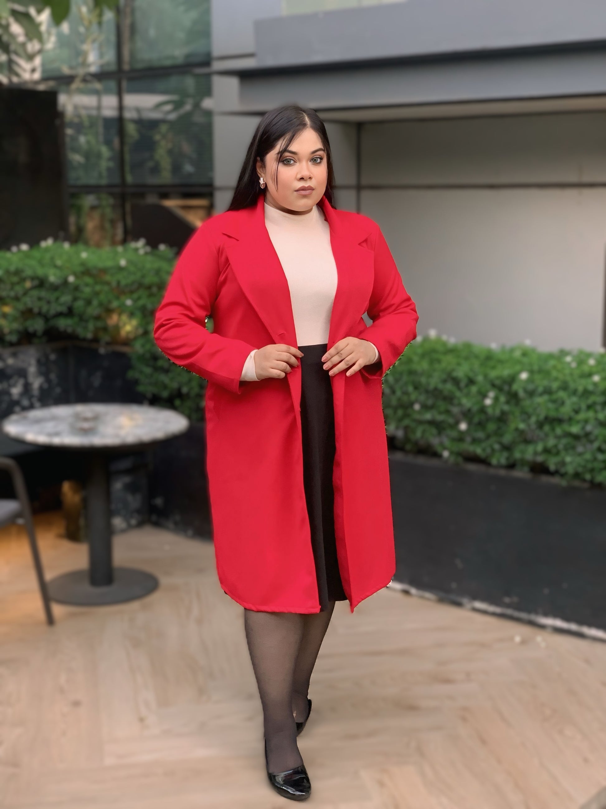Woman wearing a red coat standing outdoors with greenery and a building in the background