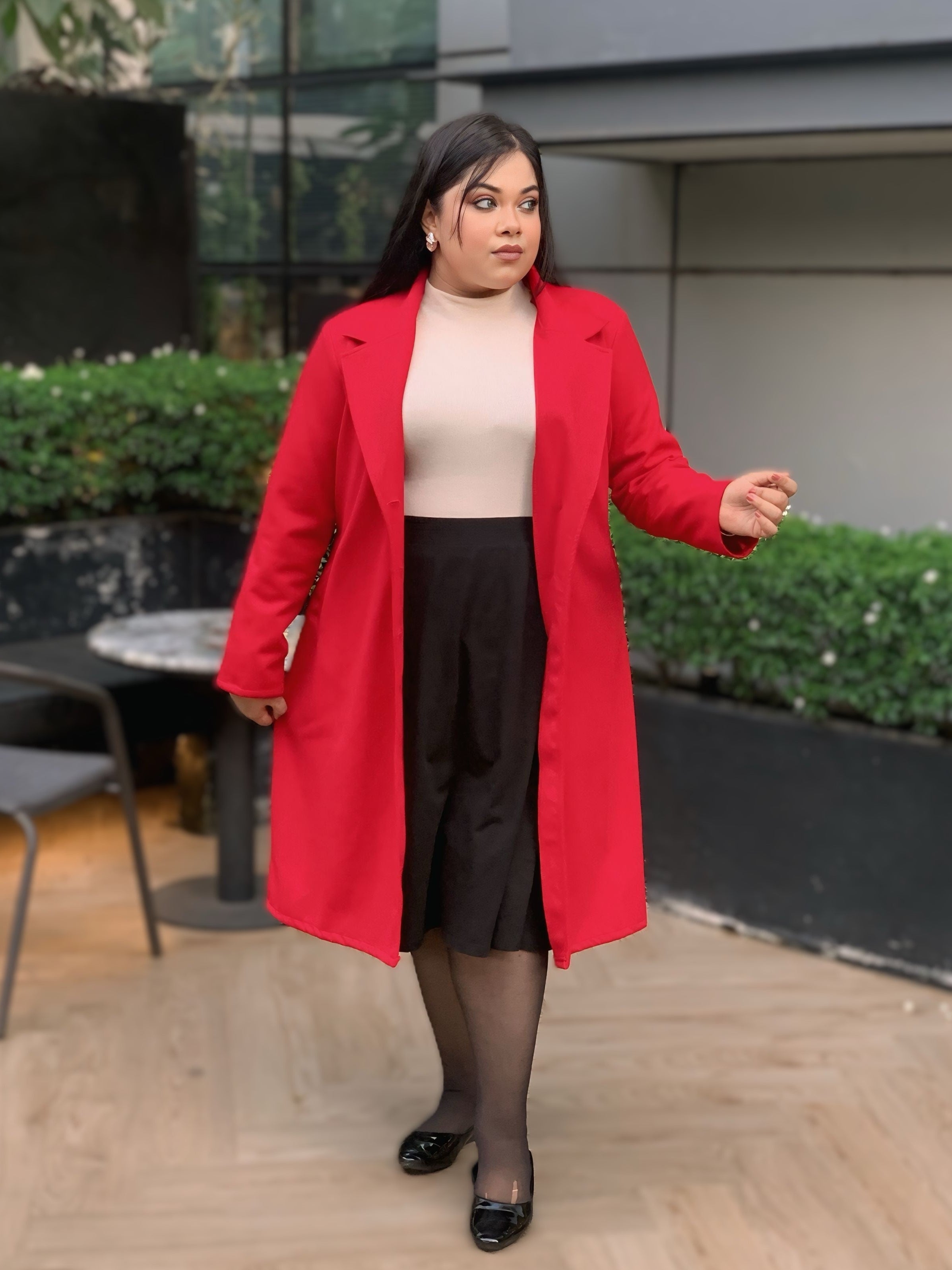 Woman wearing a red coat standing outdoors with greenery and a building in the background