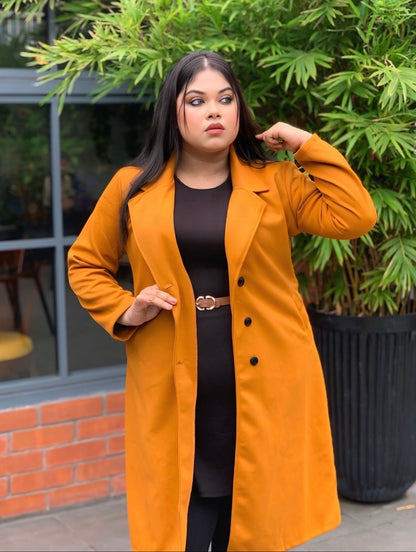 Woman wearing a mustard yellow coat standing outdoors with greenery in the background