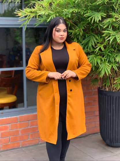 Woman wearing a mustard yellow coat standing outdoors next to a building with large windows and greenery.