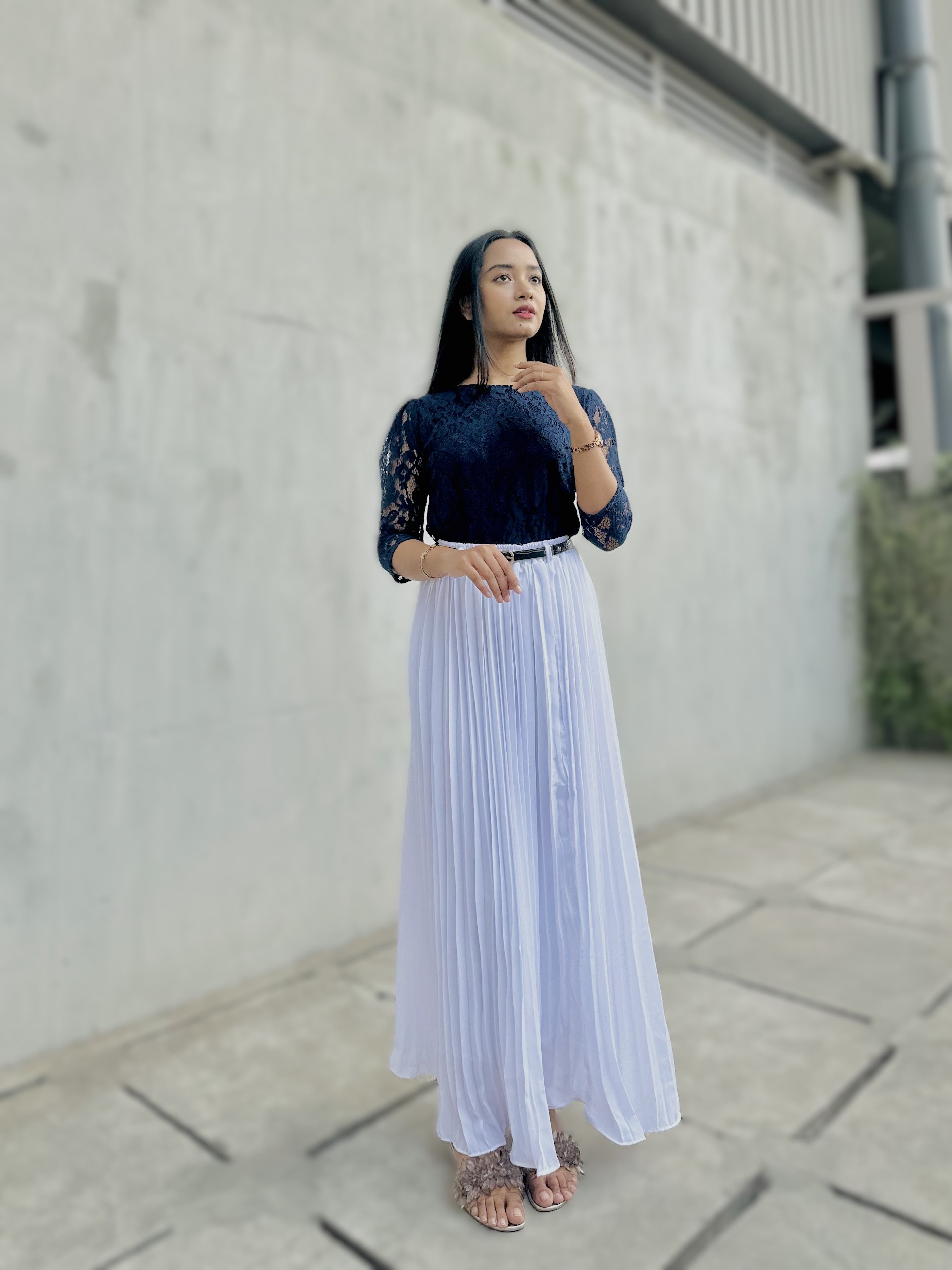 Woman wearing a black top and white pleated skirt standing against a light gray wall.