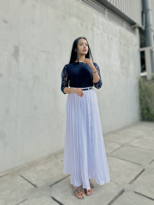 Woman wearing a black top and white pleated skirt standing against a light gray wall.