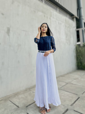 Woman wearing a navy top and white pleated skirt standing against a concrete wall.