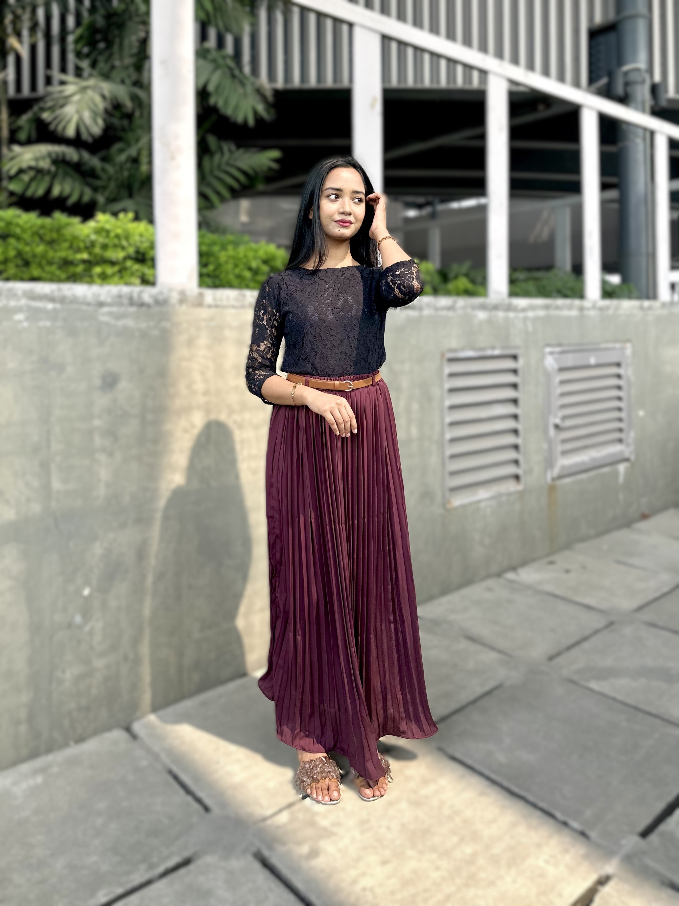 Woman in black top and purple pleated skirt standing outdoors on a concrete path.