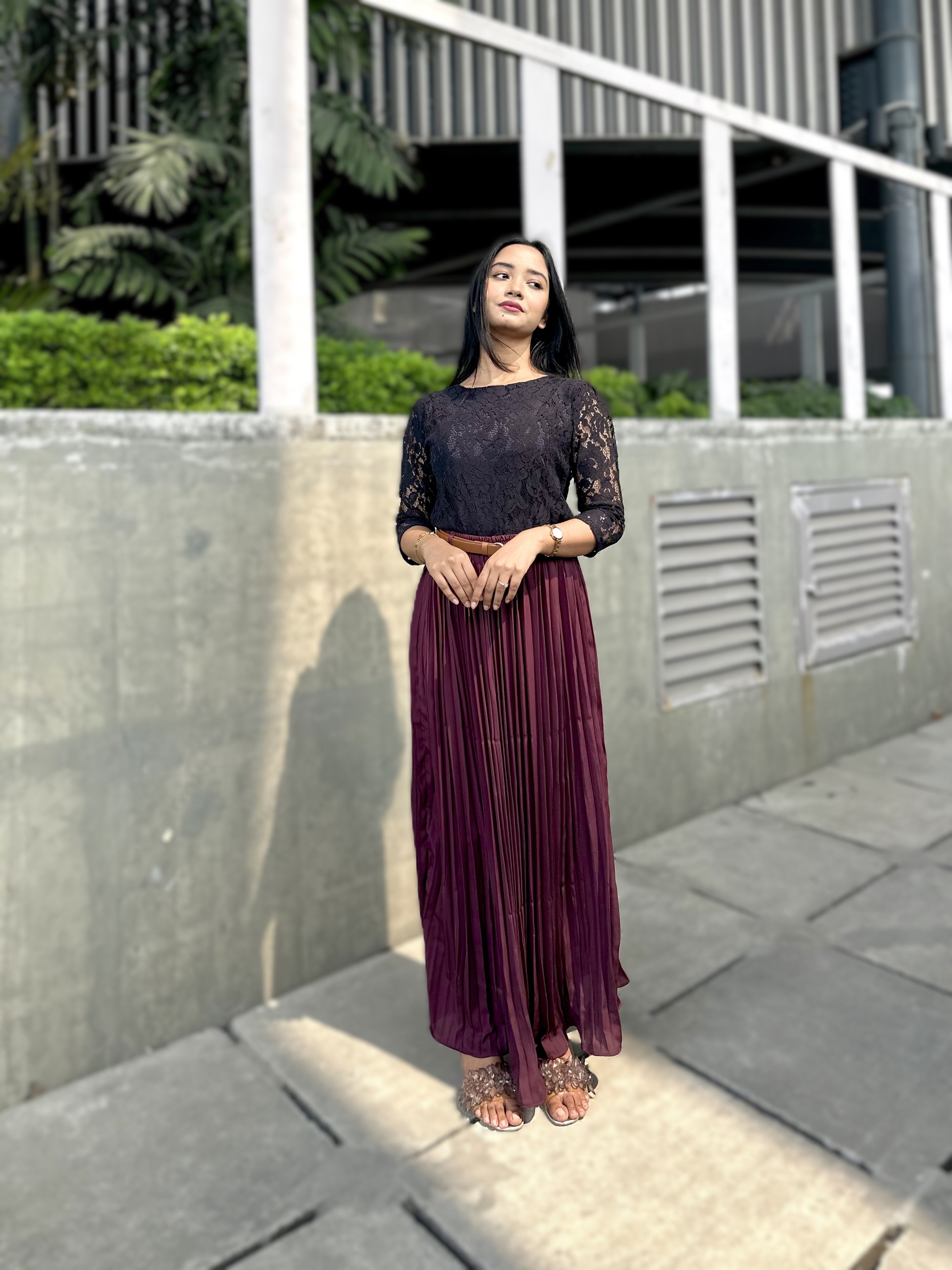 Woman in a black top and purple pleated skirt standing outdoors on a sidewalk.