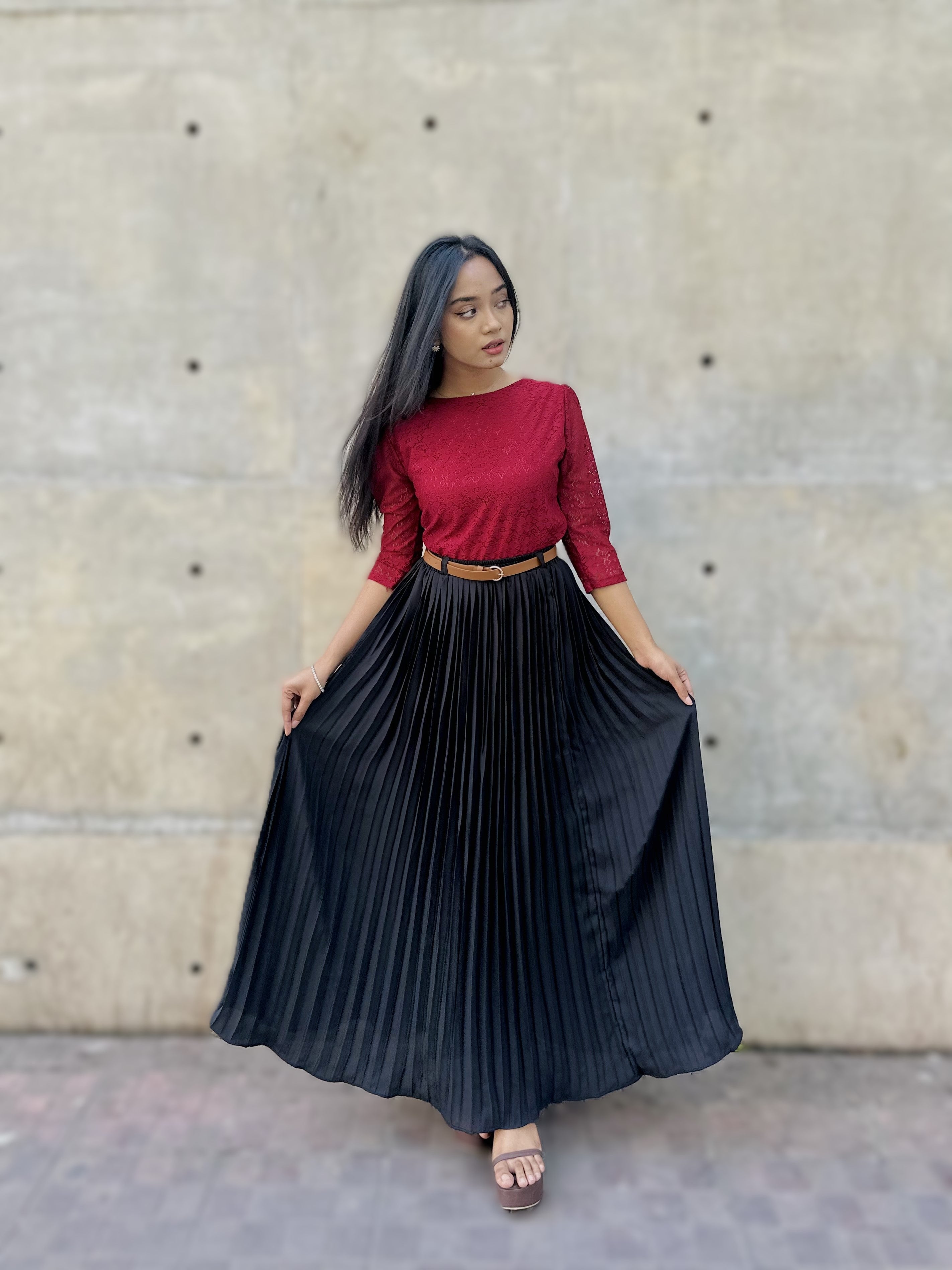 Woman wearing a red top and black pleated skirt against a concrete wall.