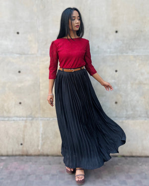 Woman wearing a red top and black pleated skirt against a beige wall.