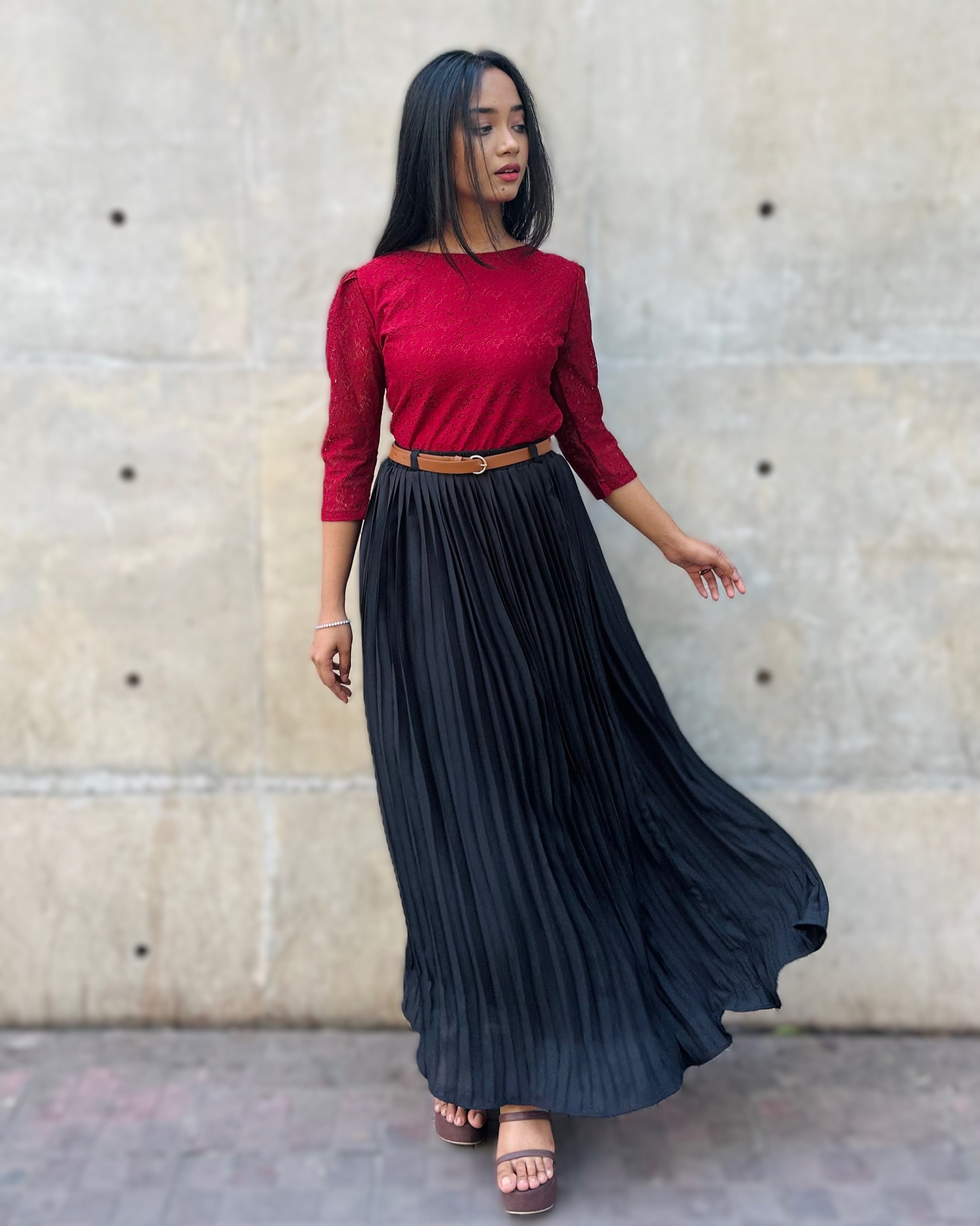 Woman wearing a red top and black pleated skirt against a beige wall.