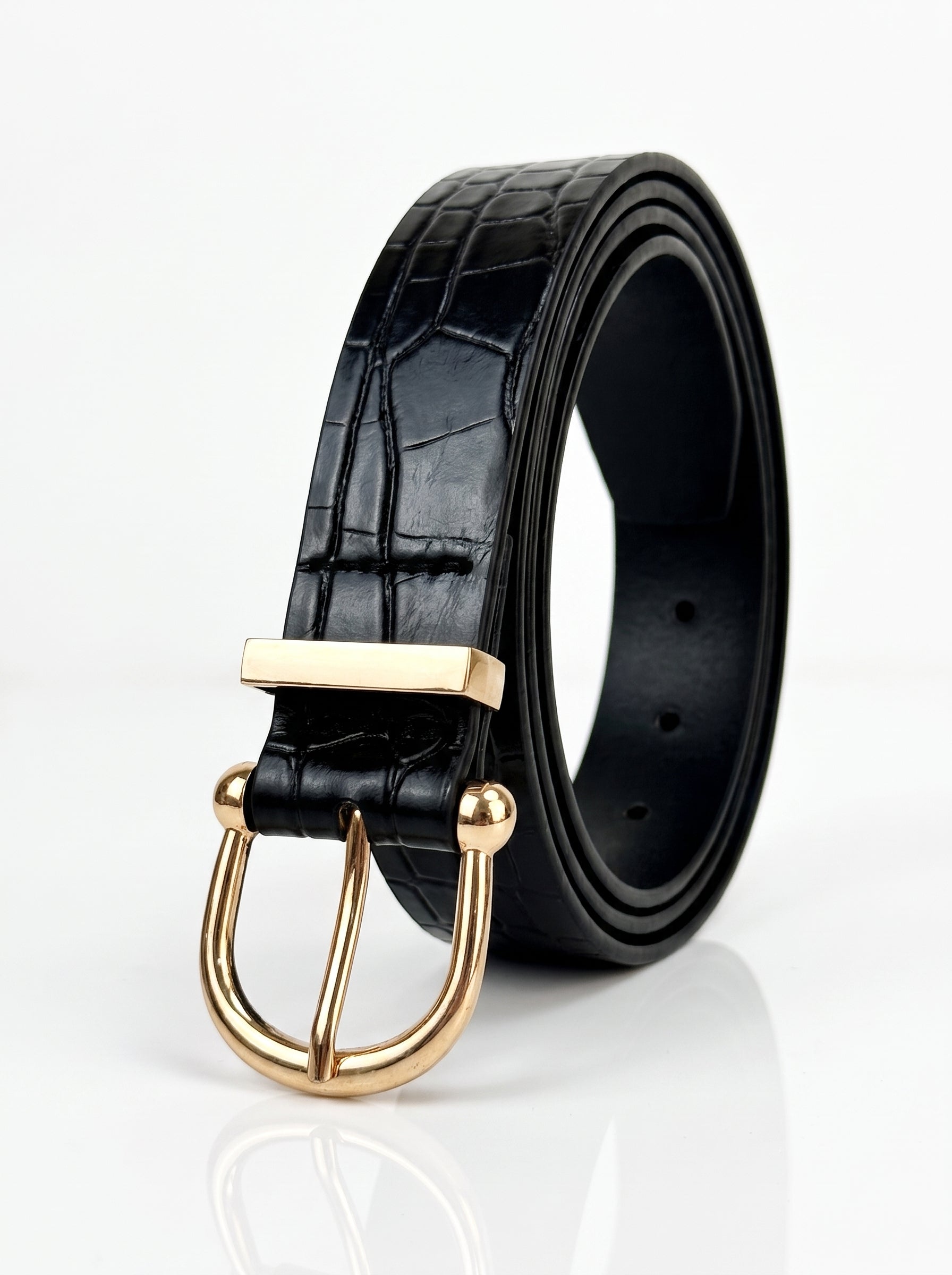 A black crocodile-embossed leather belt with a polished gold-tone metal buckle, shown coiled on a reflective white surface.