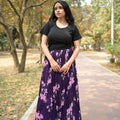 Woman in a black top and purple floral dress standing outdoors with trees and grass in the background TrendyBuzz