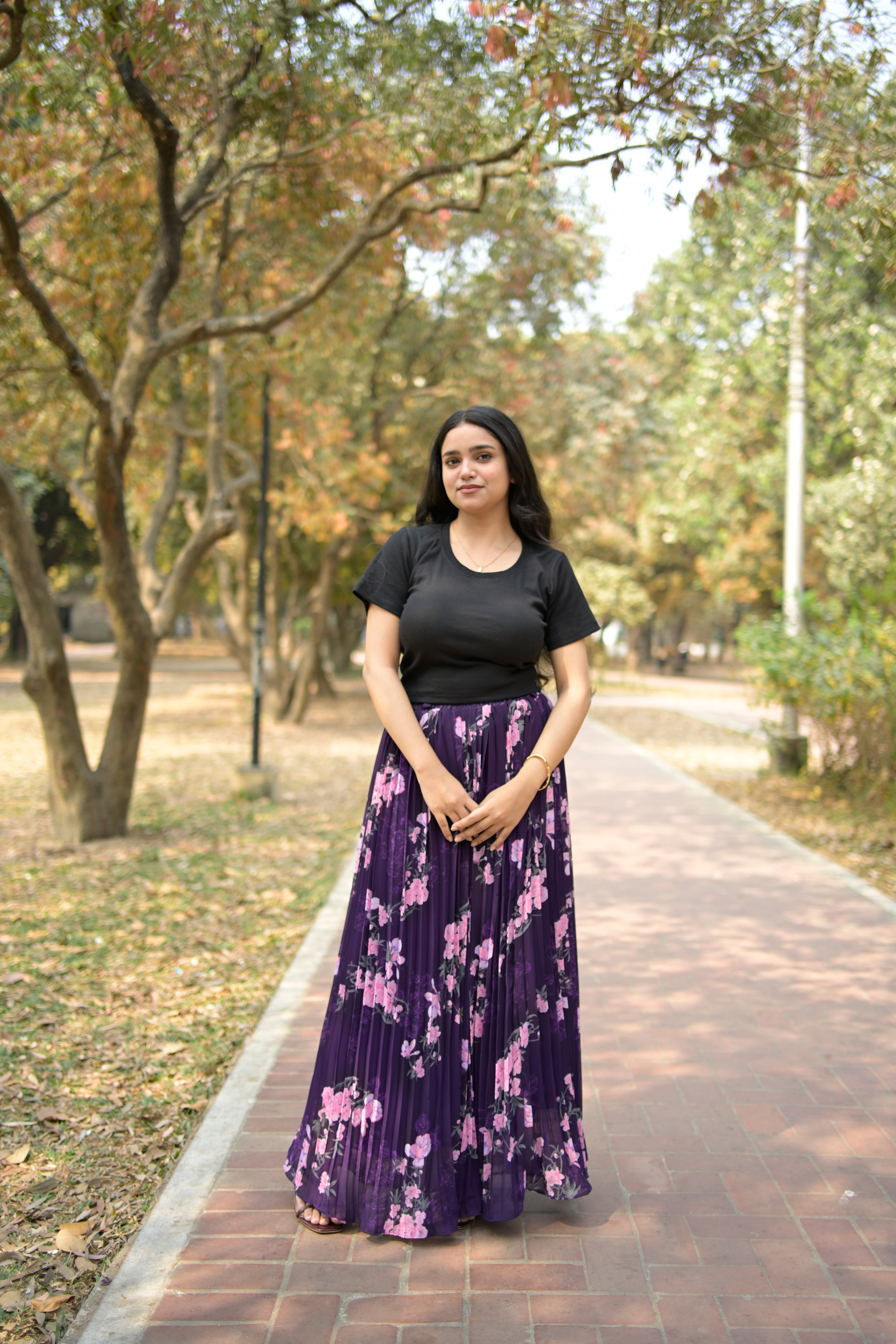 Woman in a black top and purple floral dress standing on a path with trees in the background TrendyBuzz