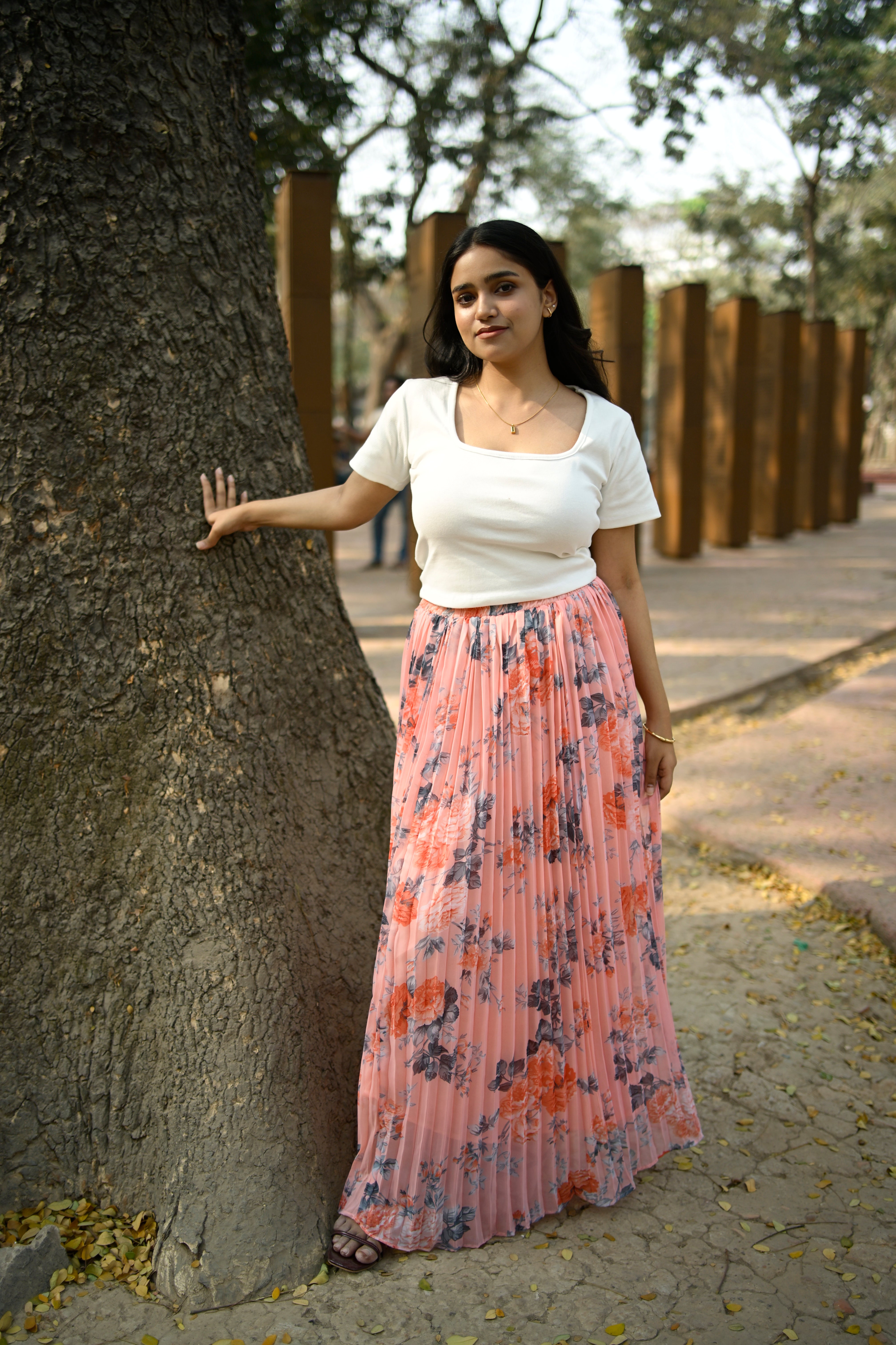 Woman in a floral dress and white blouse leaning against a tree