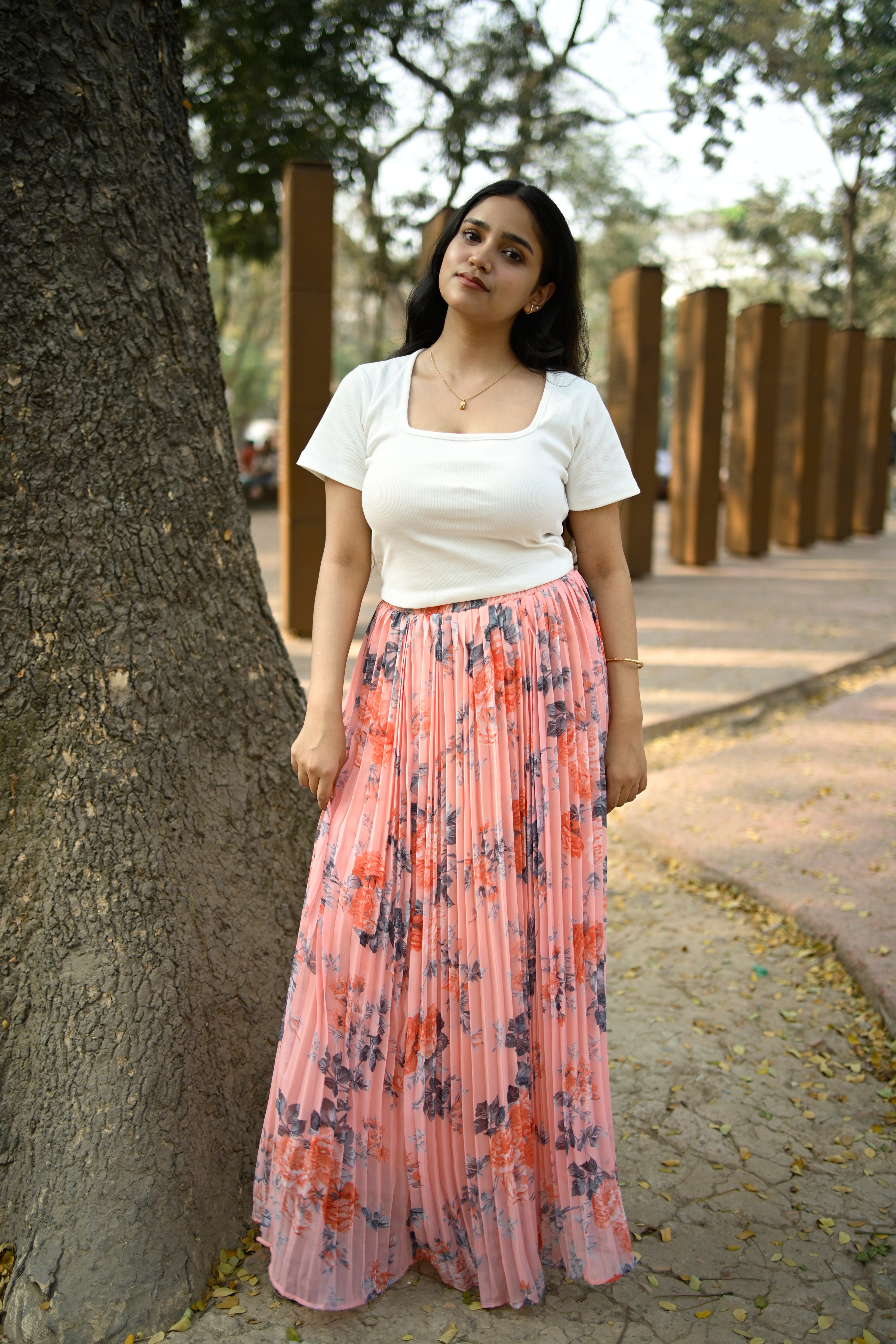 Woman in a white top and floral skirt standing next to a tree on a path