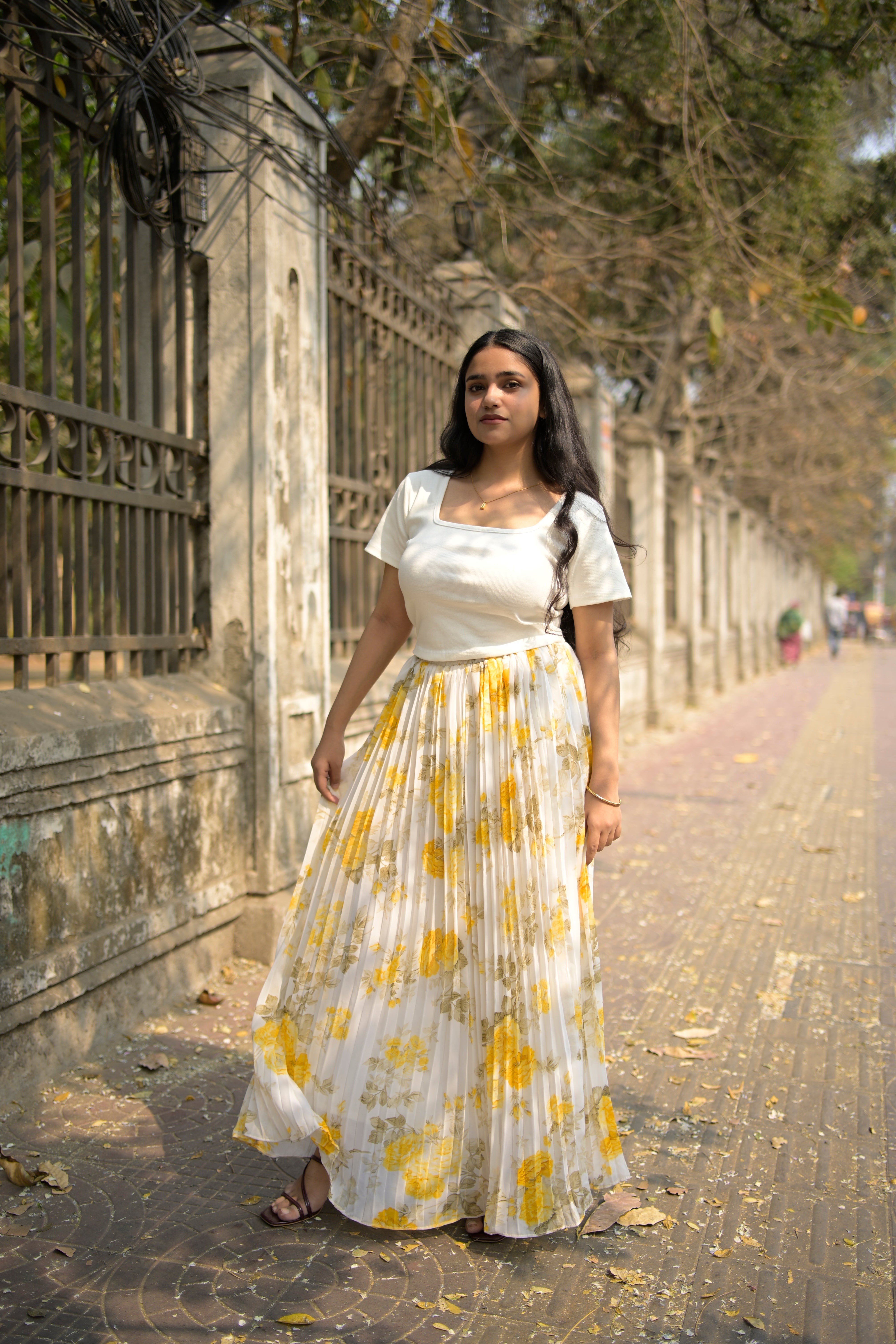 Woman in a white dress with yellow floral patterns standing on a stone pathway.