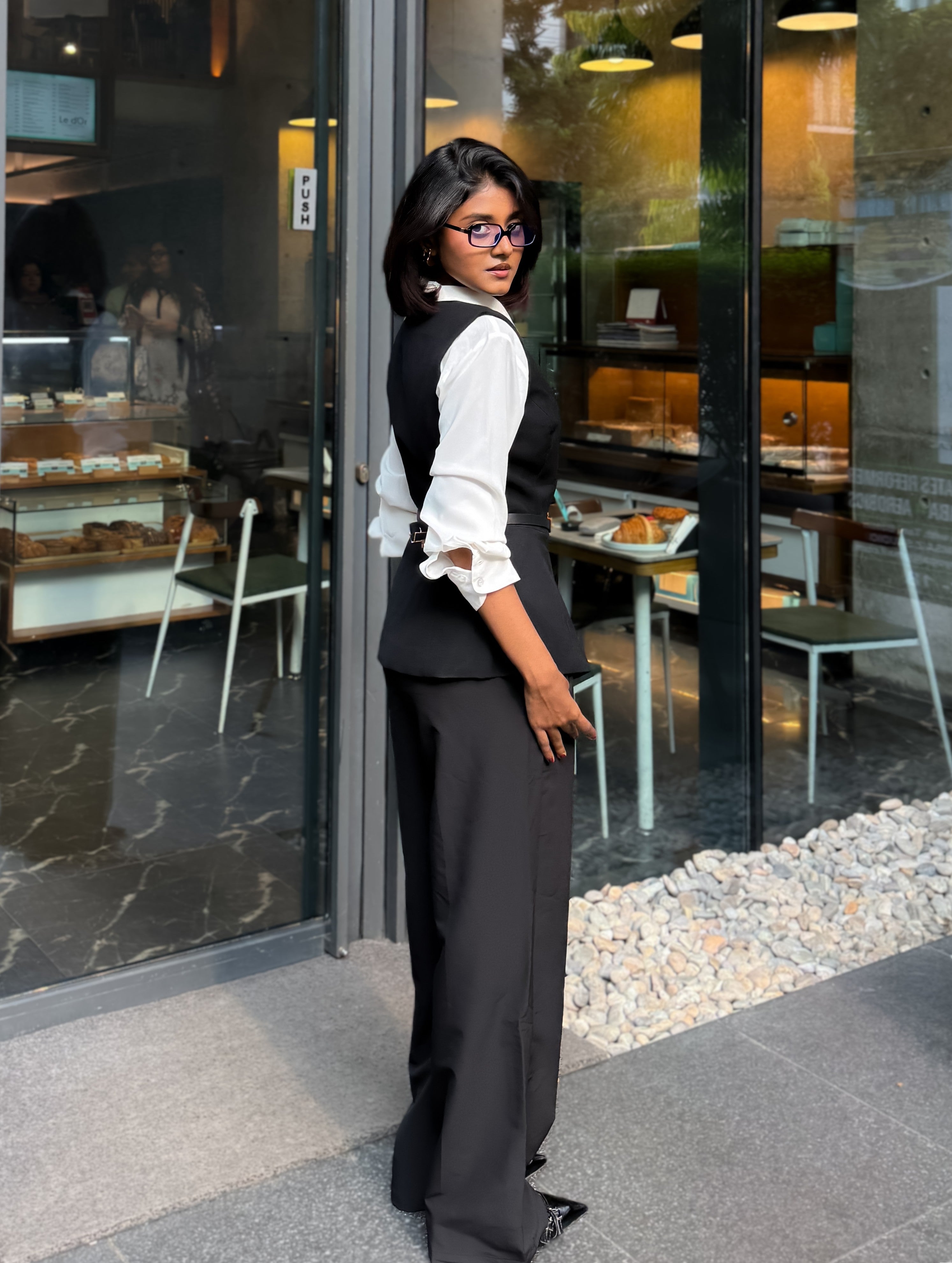 A woman wearing a black vest and wide-leg black trousers over a white long-sleeve blouse, standing in front of a glass-front cafe storefront.