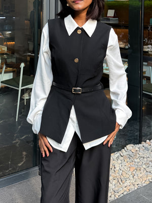 A woman wearing a black structured vest over a white collared blouse with matching black trousers, standing in front of a glass storefront.