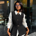 A woman wearing a black tailored vest and matching trousers over a white long-sleeve collared shirt, styled with a thin black belt and glasses, standing in front of a modern glass-front building.