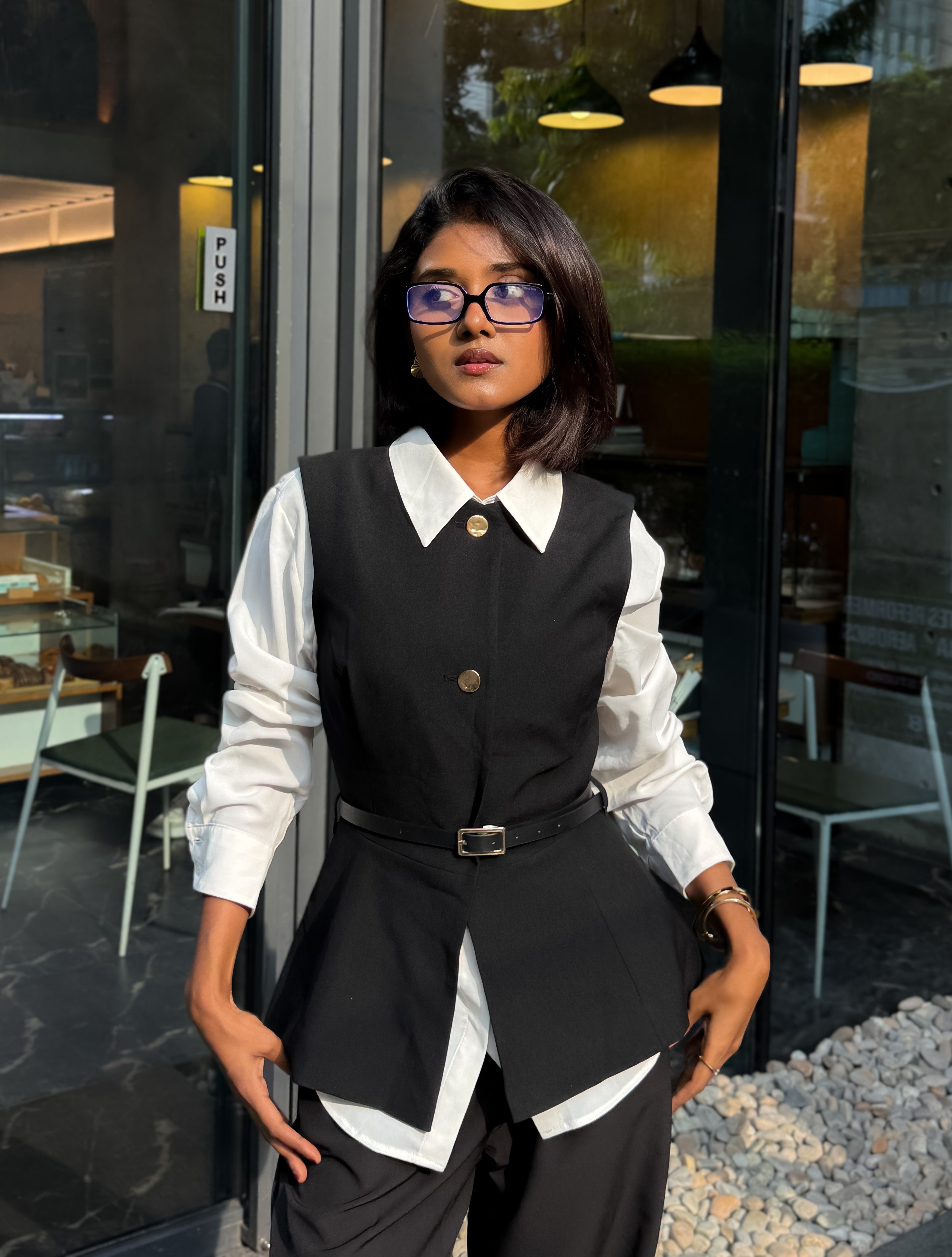 A woman wearing a black tailored vest and matching trousers over a white long-sleeve collared shirt, styled with a thin black belt and glasses, standing in front of a modern glass-front building.