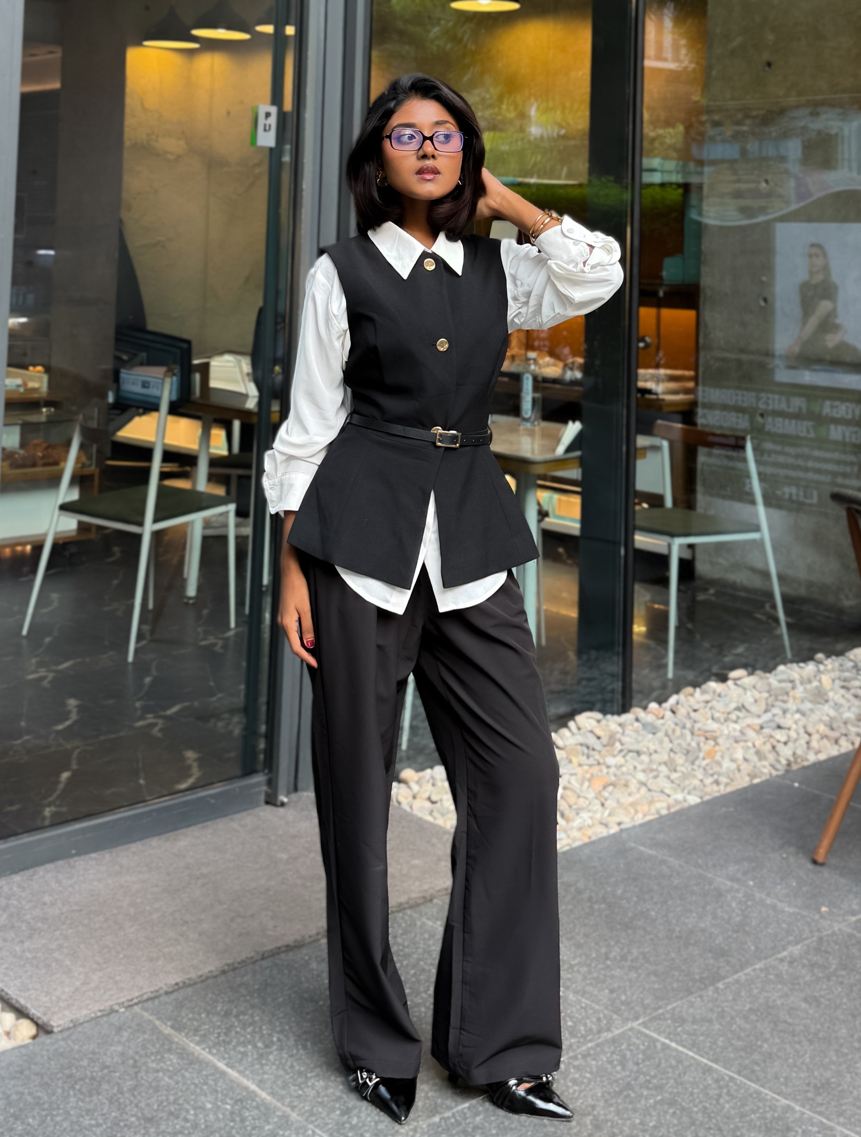 A woman models a black vest and wide-leg trousers set with a white collared shirt, standing in front of a glass storefront.