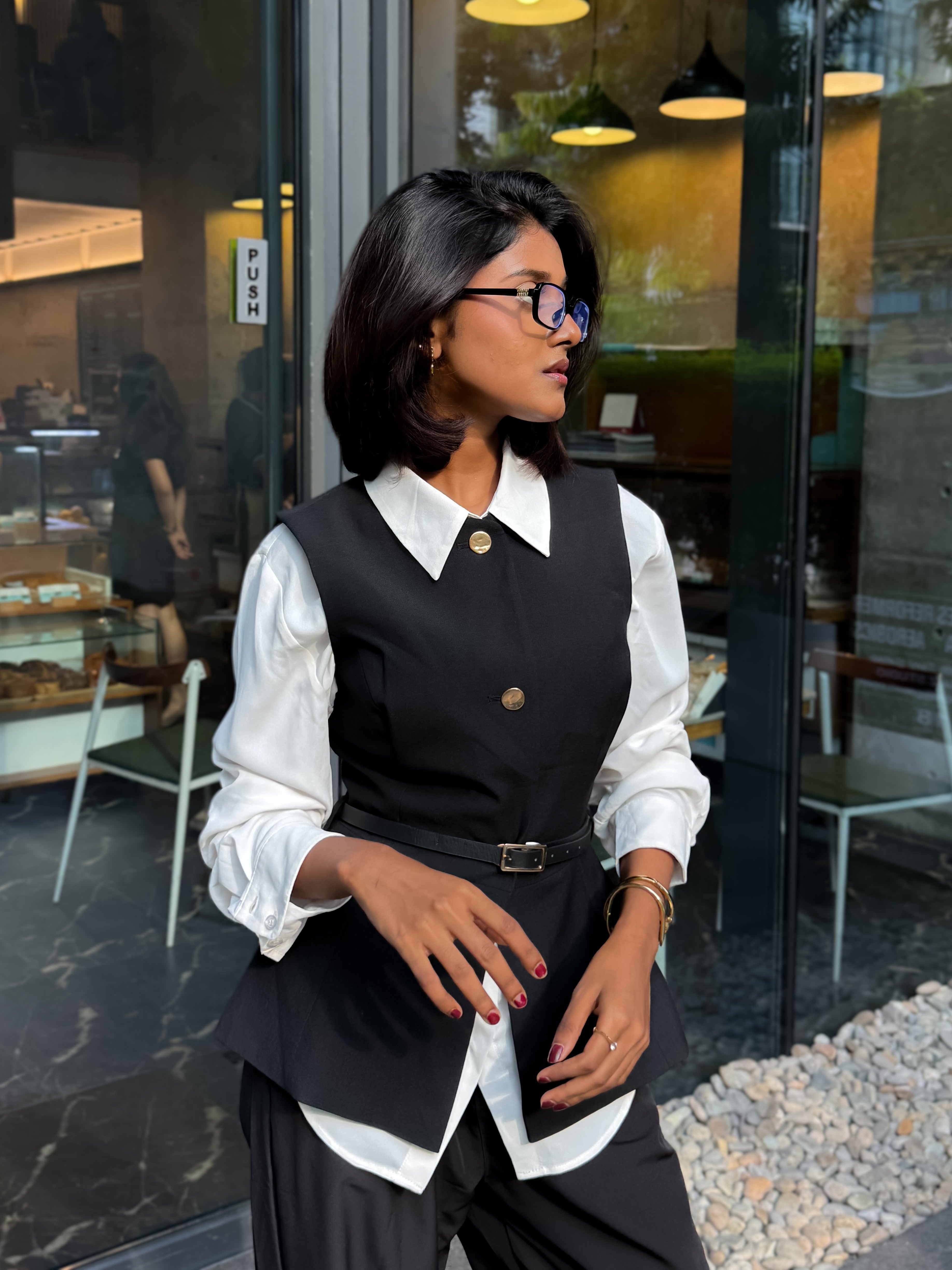 A woman wearing a black structured vest over a white collared blouse, paired with black trousers and glasses, standing in front of a glass storefront.
