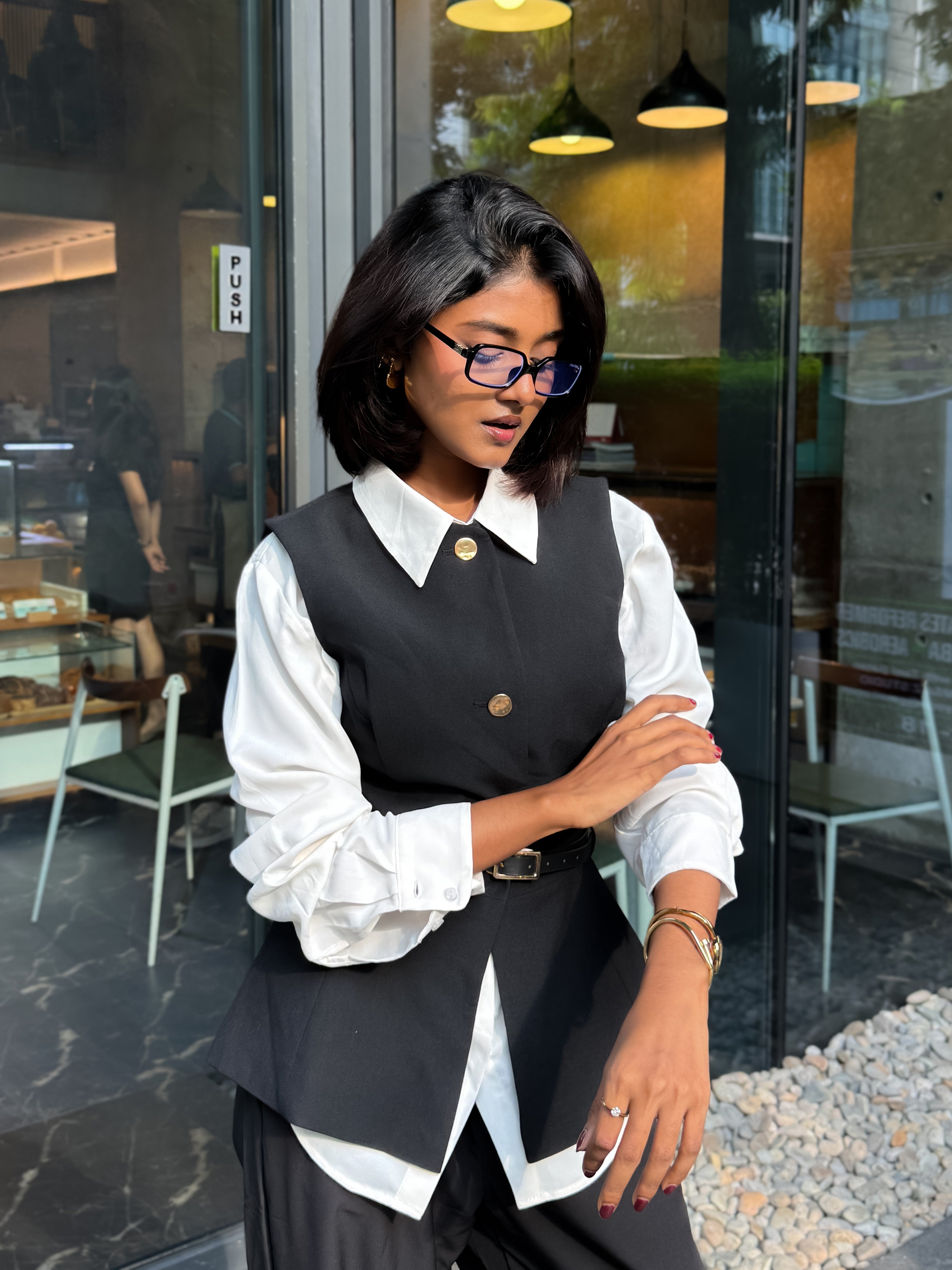 A woman wearing a black button-front vest over a white long-sleeve blouse, standing in front of a glass storefront.