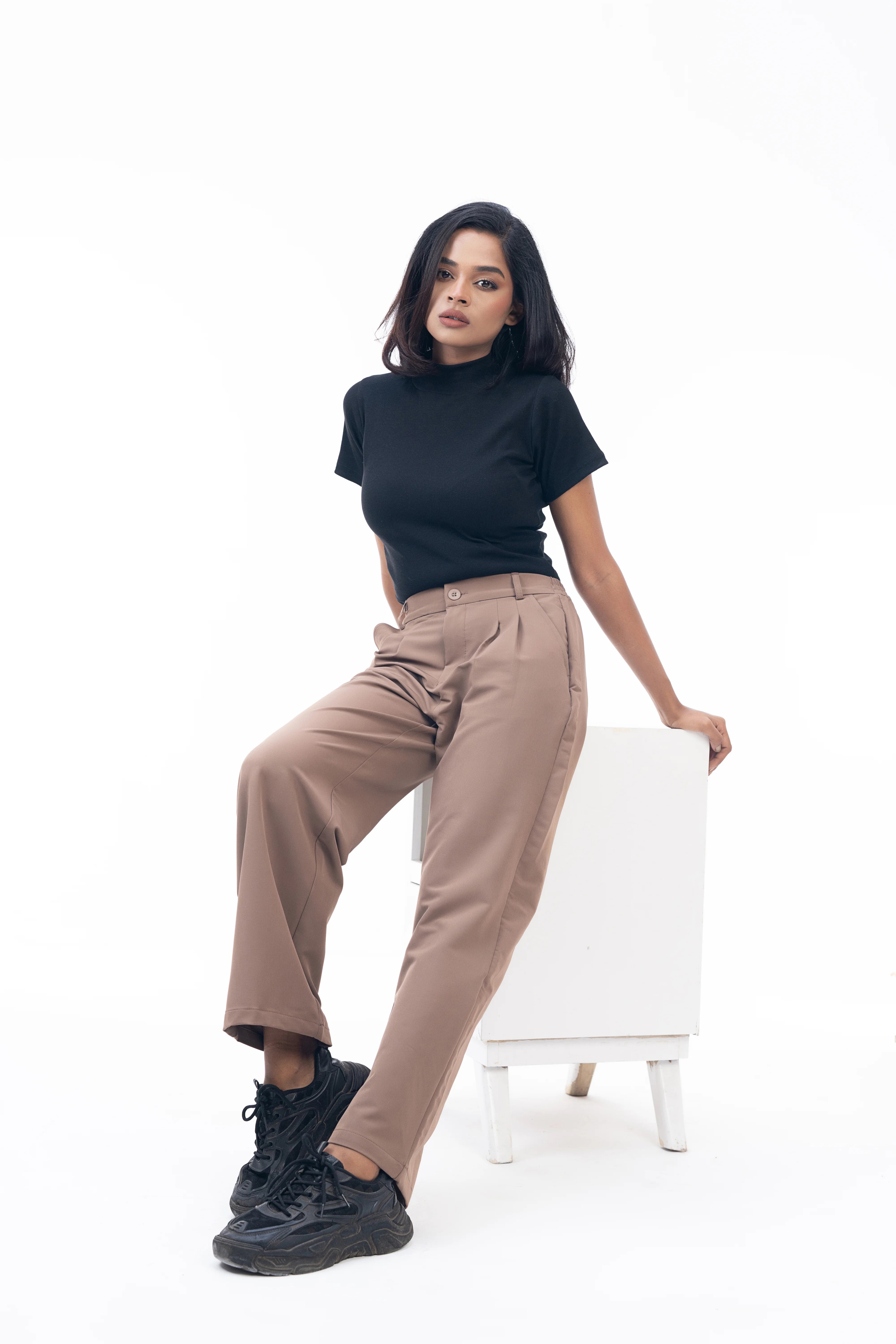 Woman sitting on a white chair wearing a black top and beige pants on a white background