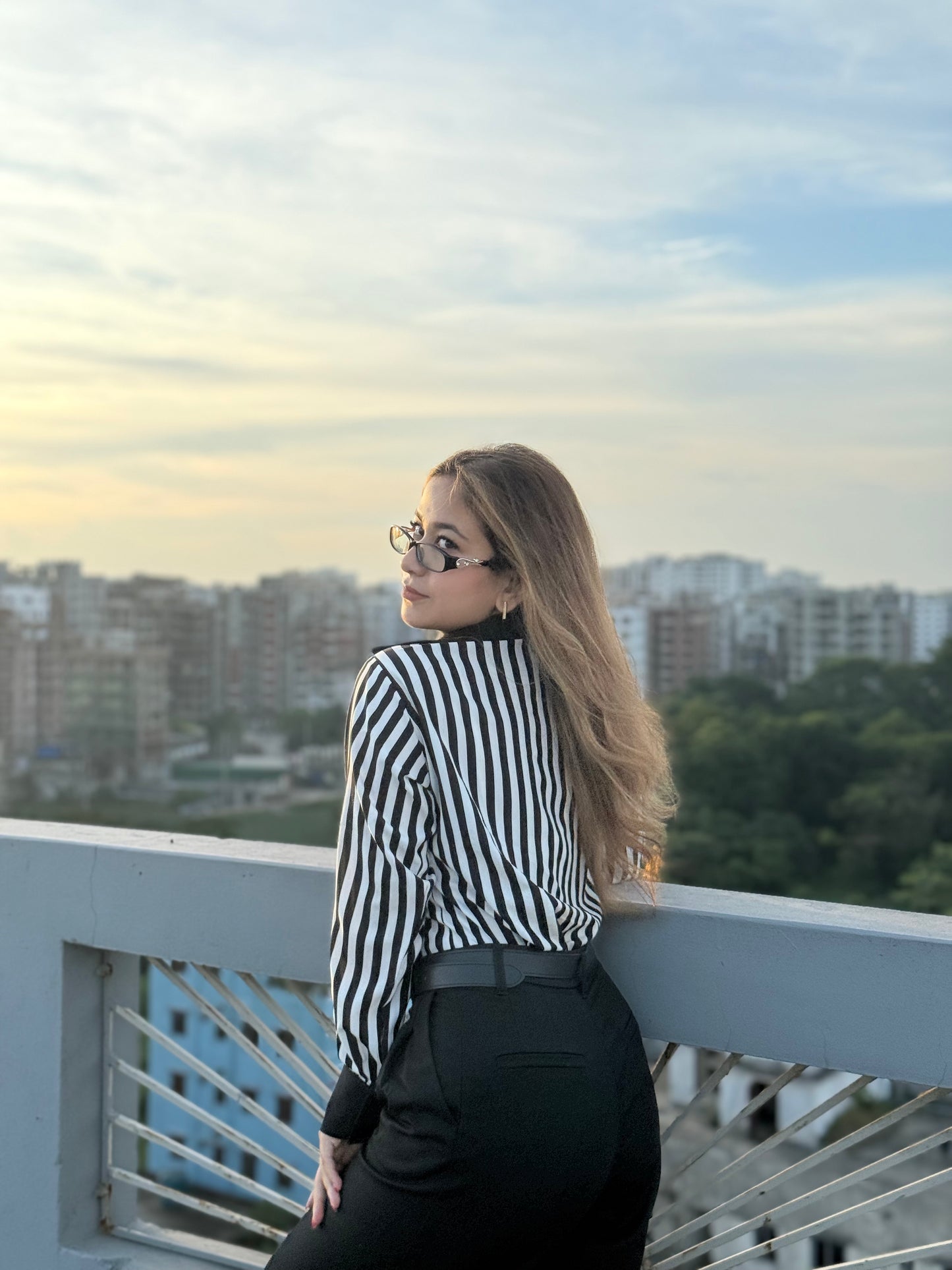 Woman in a black and white striped shirt and sunglasses standing on a balcony with a cityscape in the background.